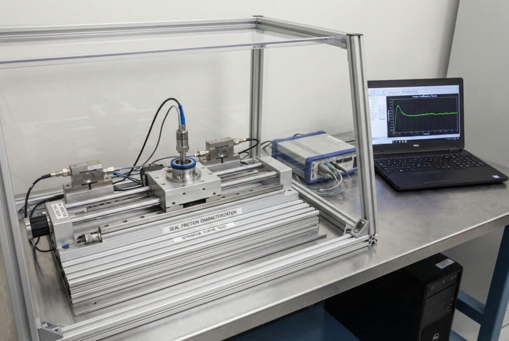 Fotogrāfija, kurā redzama laboratorijas testa iekārta blīvju berzes raksturošanai, kurā ir lineārs tribometrs caurspīdīgā korpusā, kas savienots ar datu ieguves vienību un klēpjdatoru, kurā tiek parādīts berzes koeficients reālajā laikā. Iekārta ir skaidri marķēta ar uzrakstiem "SEAL FRICTION CHARACTERIZATION" (blīvju berzes raksturošana) un "STRIBECK CURVE TEST" (Stribeka līknes tests), kas ilustrē iekārtu, ko izmanto Stribeka līkņu ģenerēšanai un berzes mērīšanai dažādos darbības apstākļos.