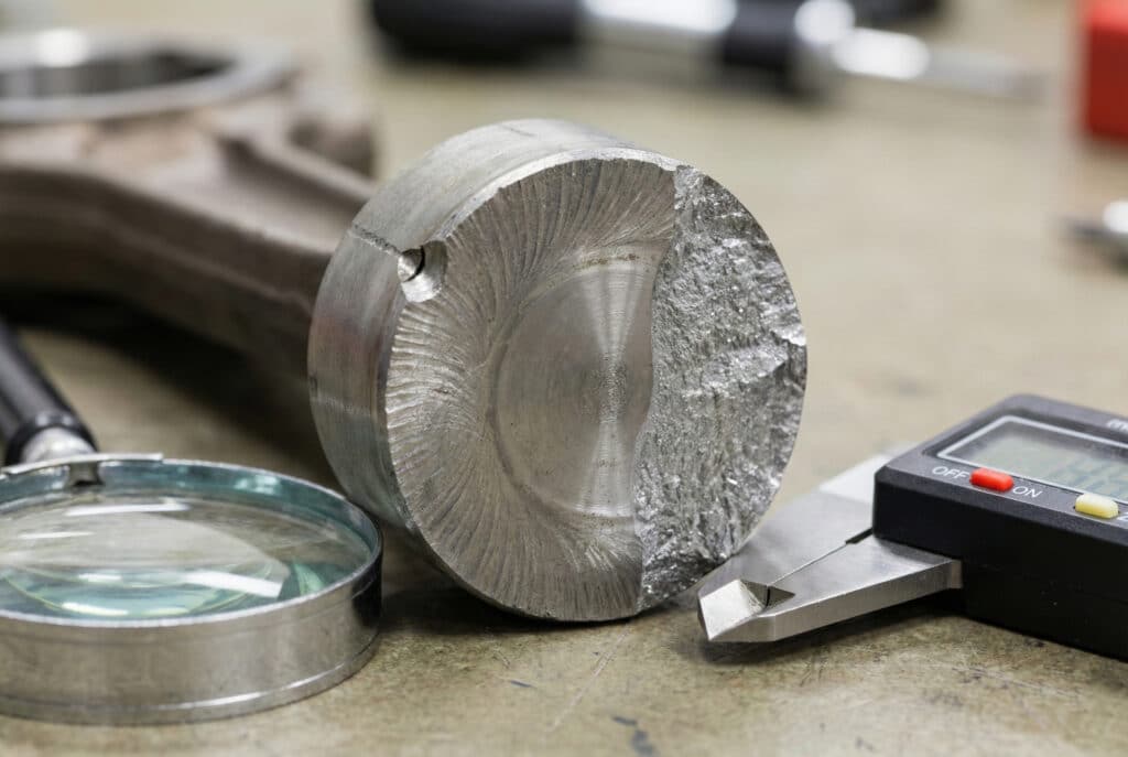 ภาพถ่ายระยะใกล้ของพื้นผิวรอยแตกของก้านลูกสูบโลหะที่หักบนโต๊ะทำงาน แสดงรอยแตกแบบชายหาดที่เป็นลักษณะเฉพาะและบริเวณรอยแตกสุดท้ายที่หยาบ อยู่ถัดจากแว่นขยายและคาลิเปอร์.