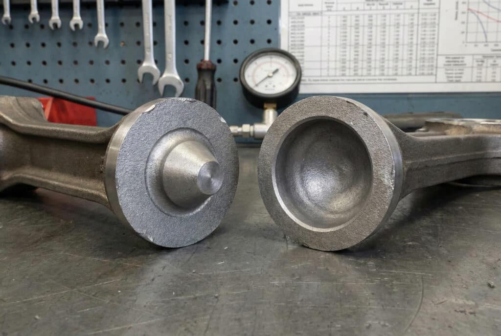 ภาพถ่ายระยะใกล้ของก้านลูกสูบโลหะที่แตกเป็นสองชิ้นบนโต๊ะทำงานในโรงงาน ชัดเจนให้เห็นลักษณะการแตกแบบถ้วยและกรวย ซึ่งเป็นลักษณะเฉพาะของการแตกหักเนื่องจากแรงดึงเกินขีดจำกัดอันเกิดจากการรับน้ำหนักเกิน.